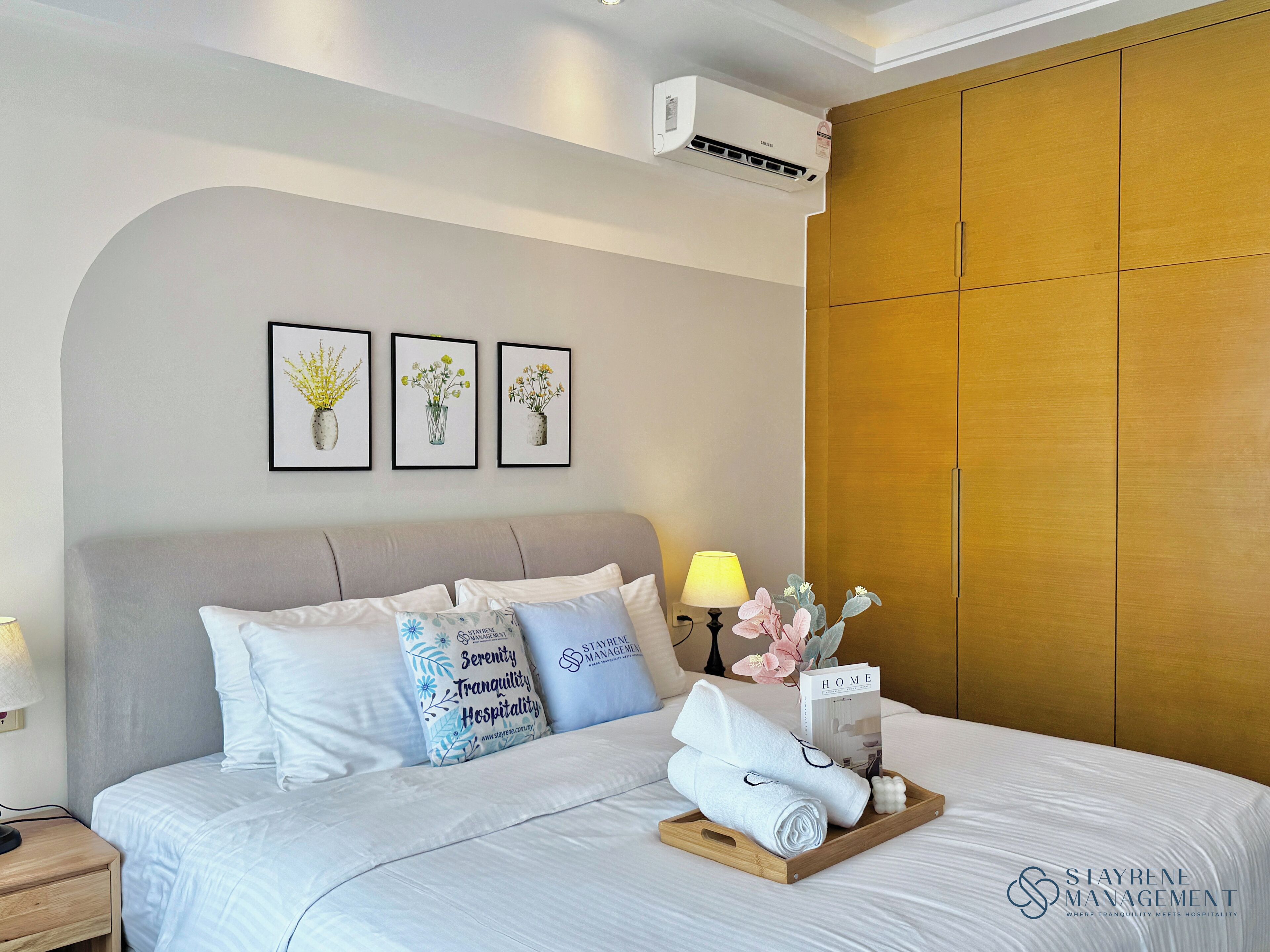 A neatly made bed with white linens, a tray with towels, and a bedside table with a lamp. There are framed floral pictures on the wall, a wooden wardrobe, and an air conditioning unit above the bed.