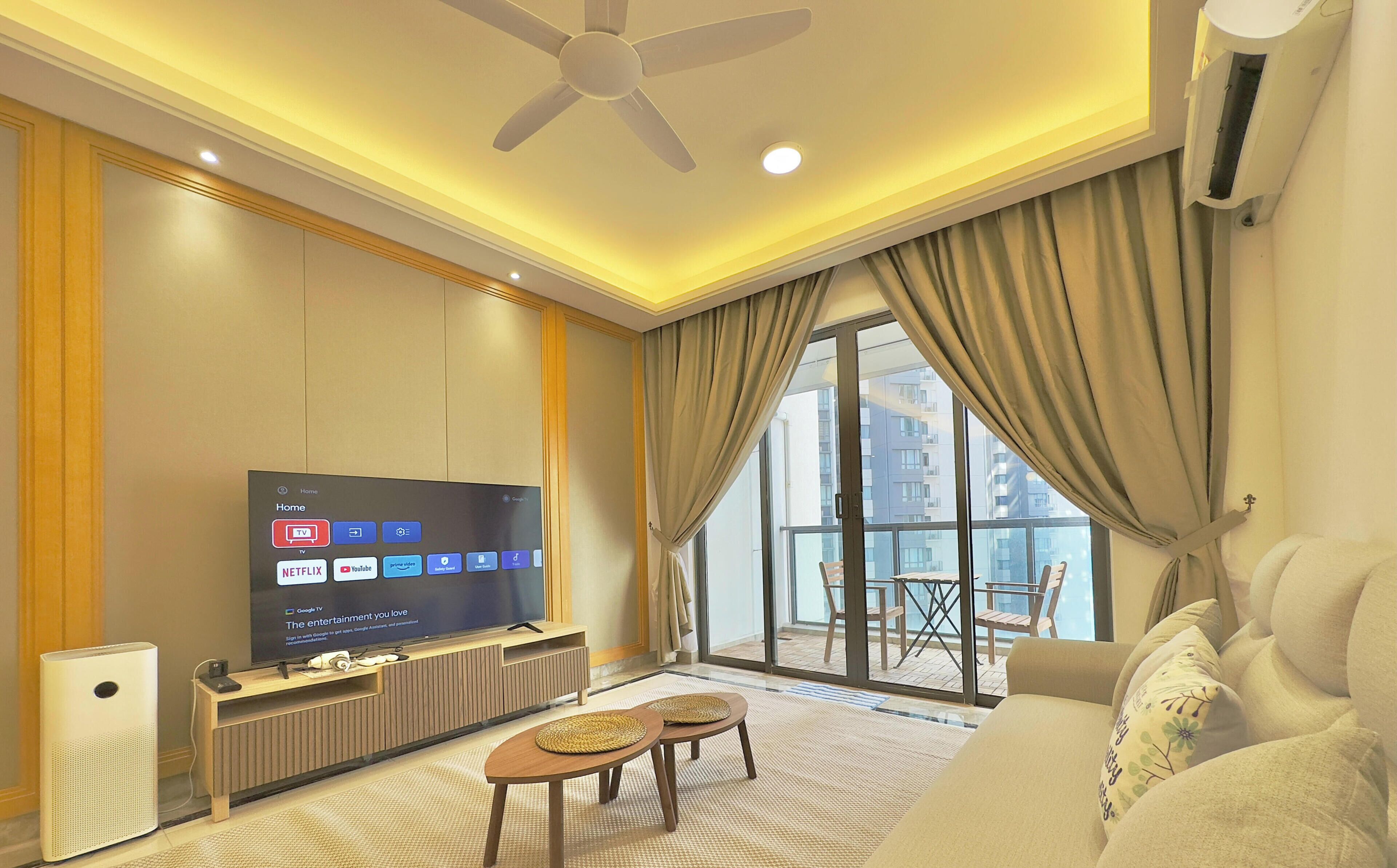 A modern hotel room with a flat-screen TV, a ceiling fan, a sofa, a small table, and a view of the city through large windows.