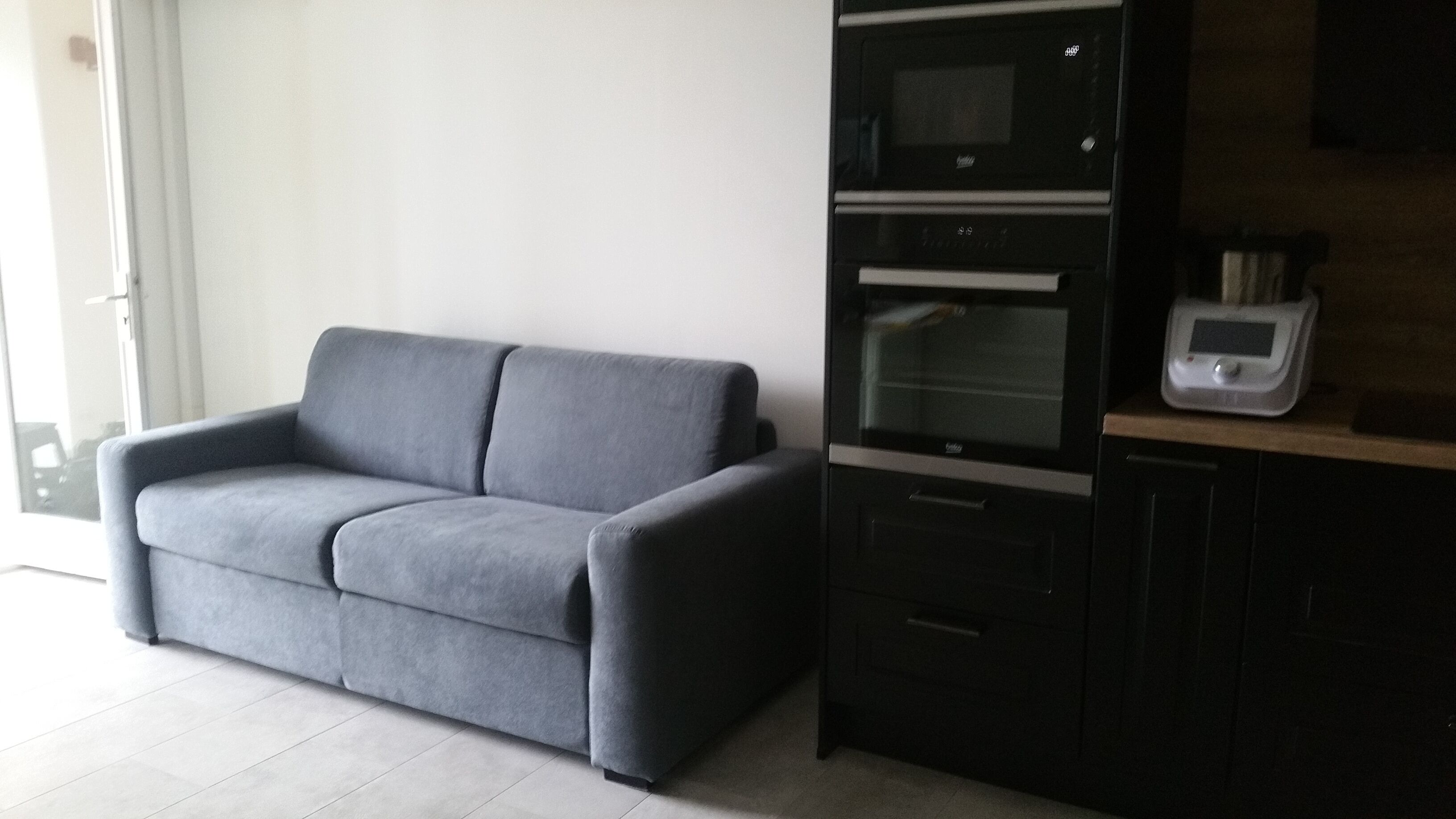 A modern kitchen with a grey sofa, built-in oven, microwave, and a small countertop with a scale.