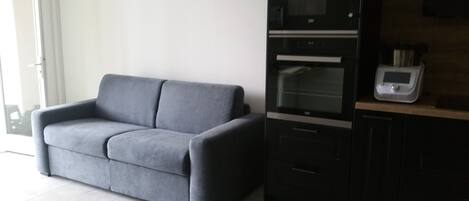 A modern kitchen with a grey sofa, built-in oven, microwave, and a small countertop with a scale.