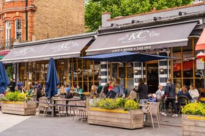 Un café al aire libre con mesas y sillas, un edificio de ladrillo al fondo y un cartel que dice KC Crêperie.