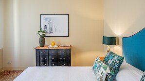 Habitació d'hotel amb un llit, una petita taula de nit, un quadre al mur, una làmpada i un coixí decoratiu.