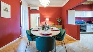 Zona de dinar amb taula rodona preparada per a sis, amb cadires color turquesa al voltant. Hi ha una làmpada penjant sobre la taula, i es veu una cuina al fons.