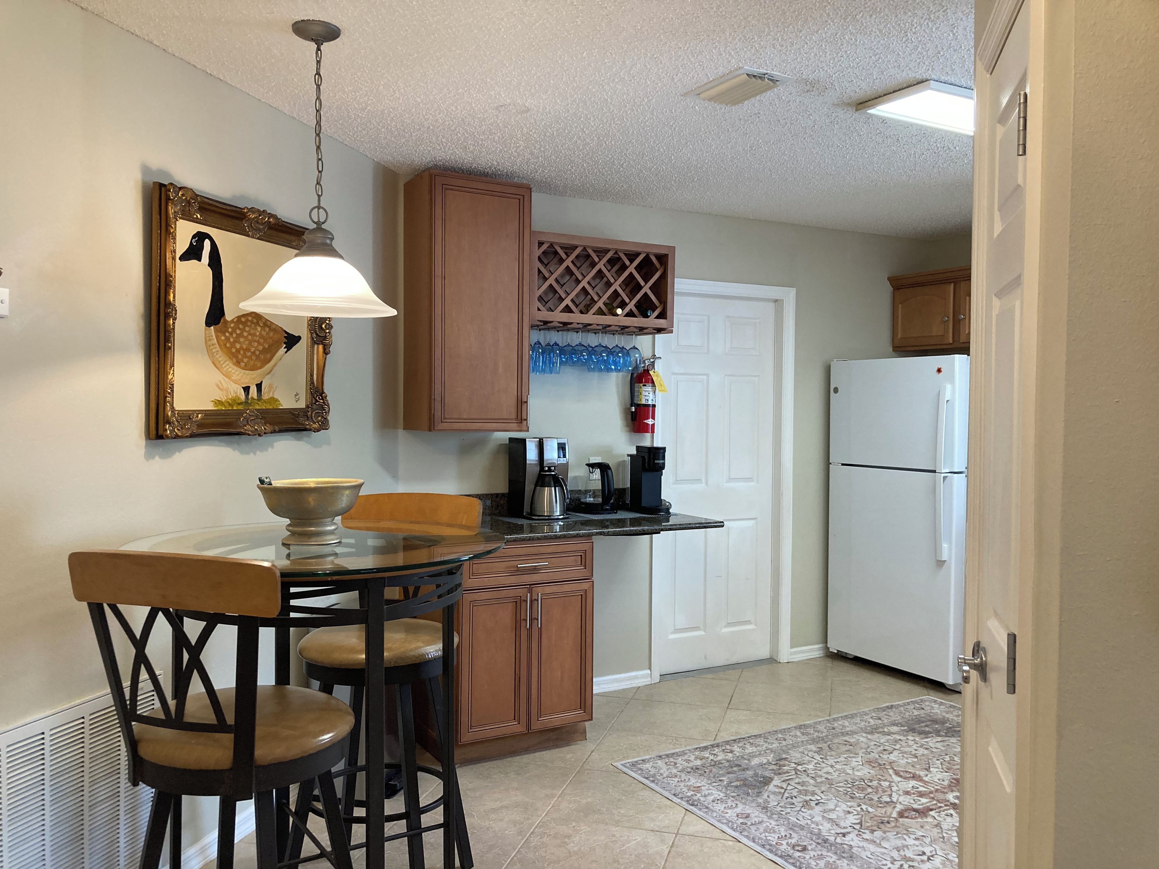 A kitchen with a dining table, chairs, a coffee maker, a refrigerator, and a fire extinguisher.