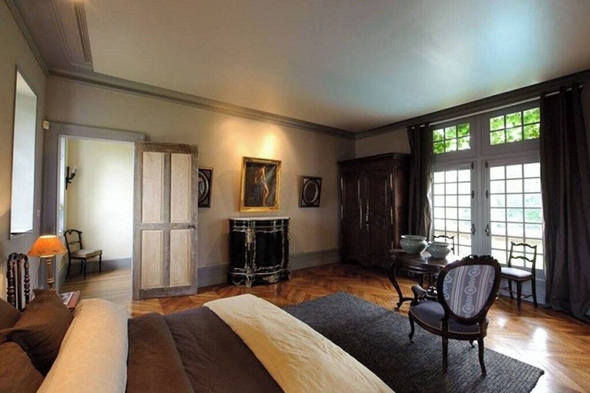 Een slaapkamer met een groot bed, een stoel, een tafel en een raam met gordijnen.