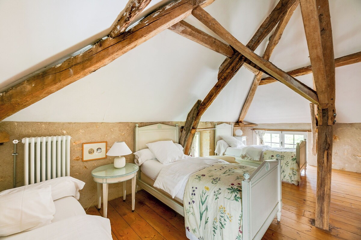 Een slaapkamer met een schuine kap, houten balken, een bed met een bloemenbedekking, een nachtkastje met een lamp en een radiator.