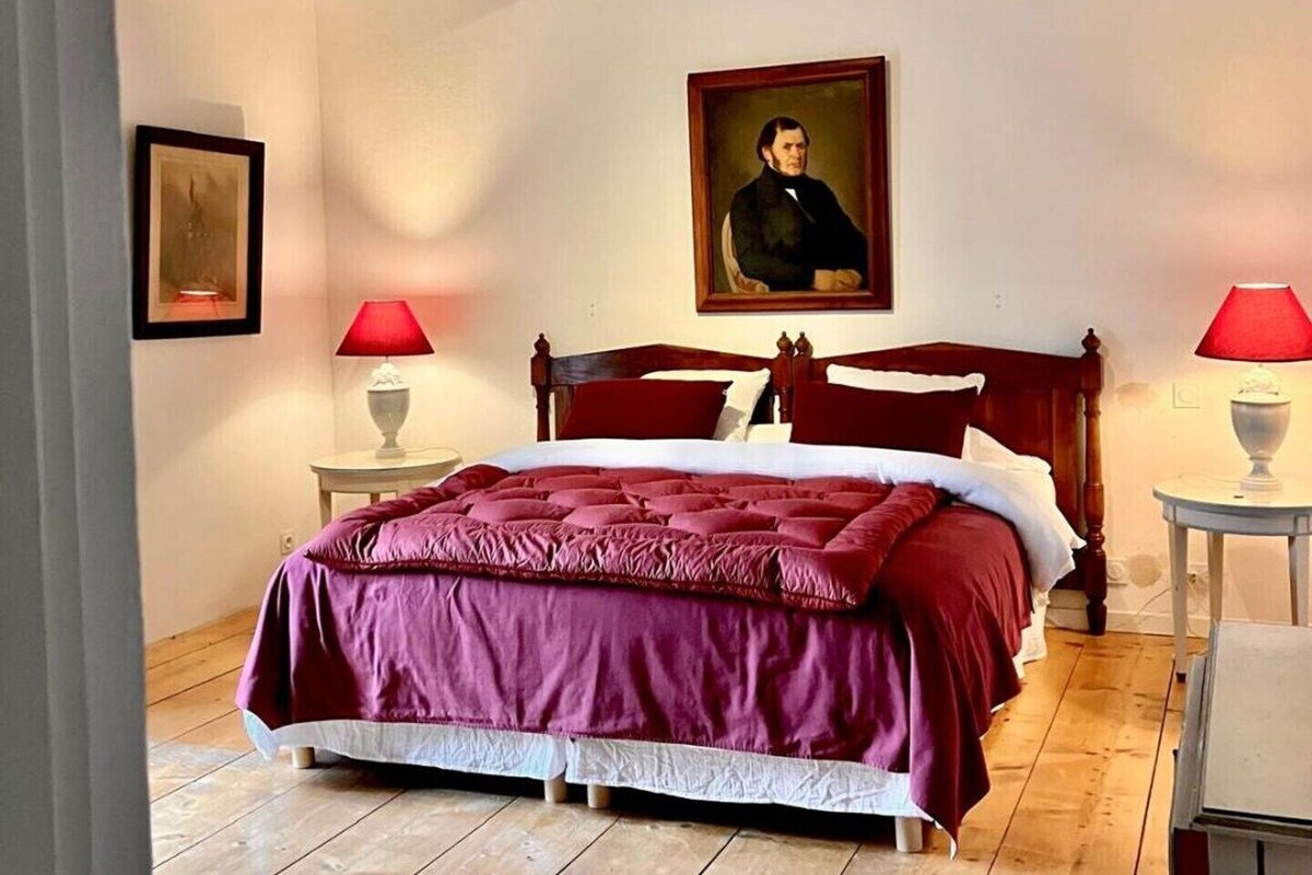 Een slaapkamer met een houten plafond, een groot bed met een rood-witte dekbedovertrek, twee nachtkastjes met lampen en een ingelijst portret aan de muur.