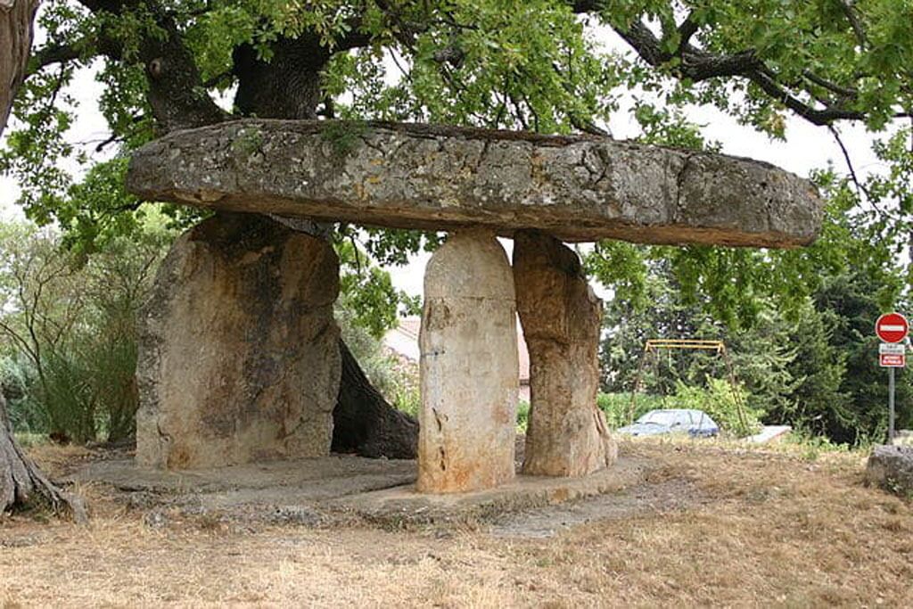 A stone structure supported by large tree trunks, surrounded by dry grass and trees.