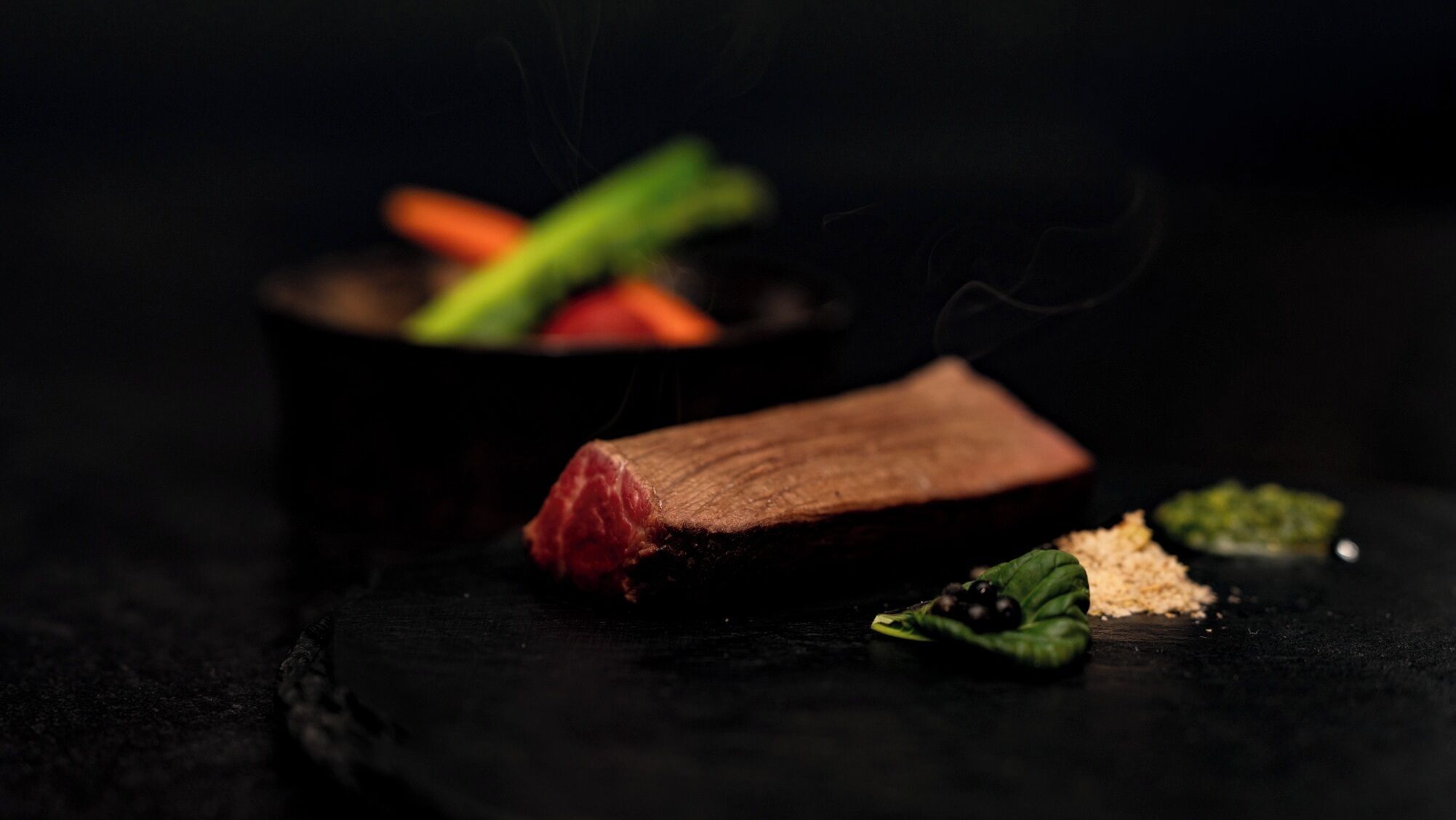 Un bife con verduras y un recipiente pequeño sobre una superficie oscura.