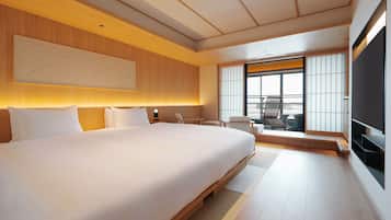 A modern hotel room with a large bed, a seating area with a chair and table, a television, and a view through the window.