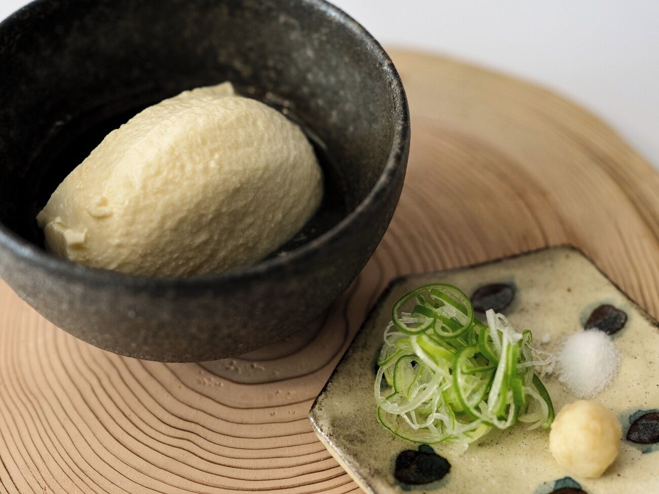A bowl of ice cream with a scoop of vanilla, a small dish with green onions, salt, and a small round item on a wooden surface.