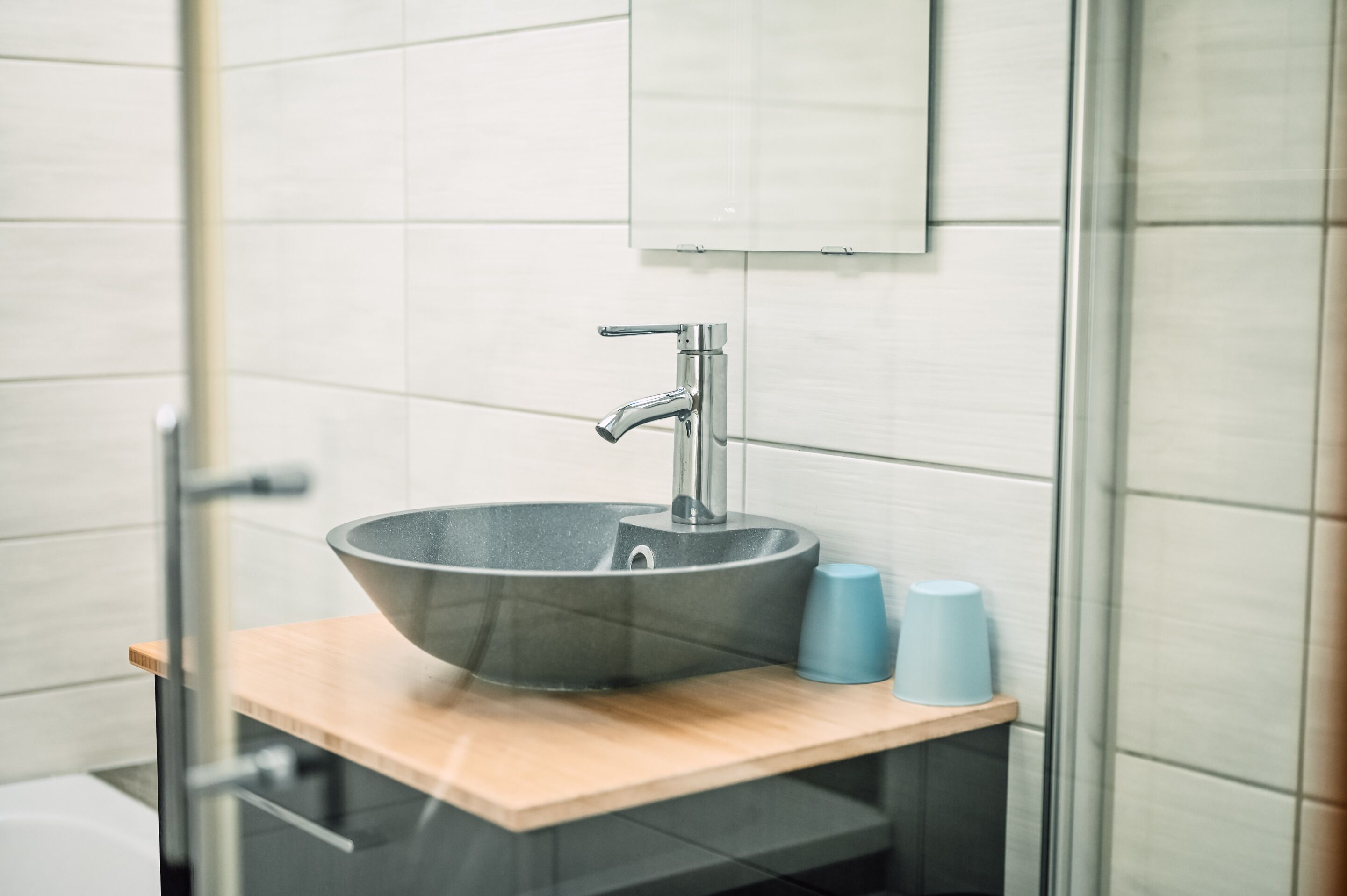 A modern bathroom with a large, round, dark-colored sink, a single faucet, and a mirror above it. There are two blue cups on the sink's edge.