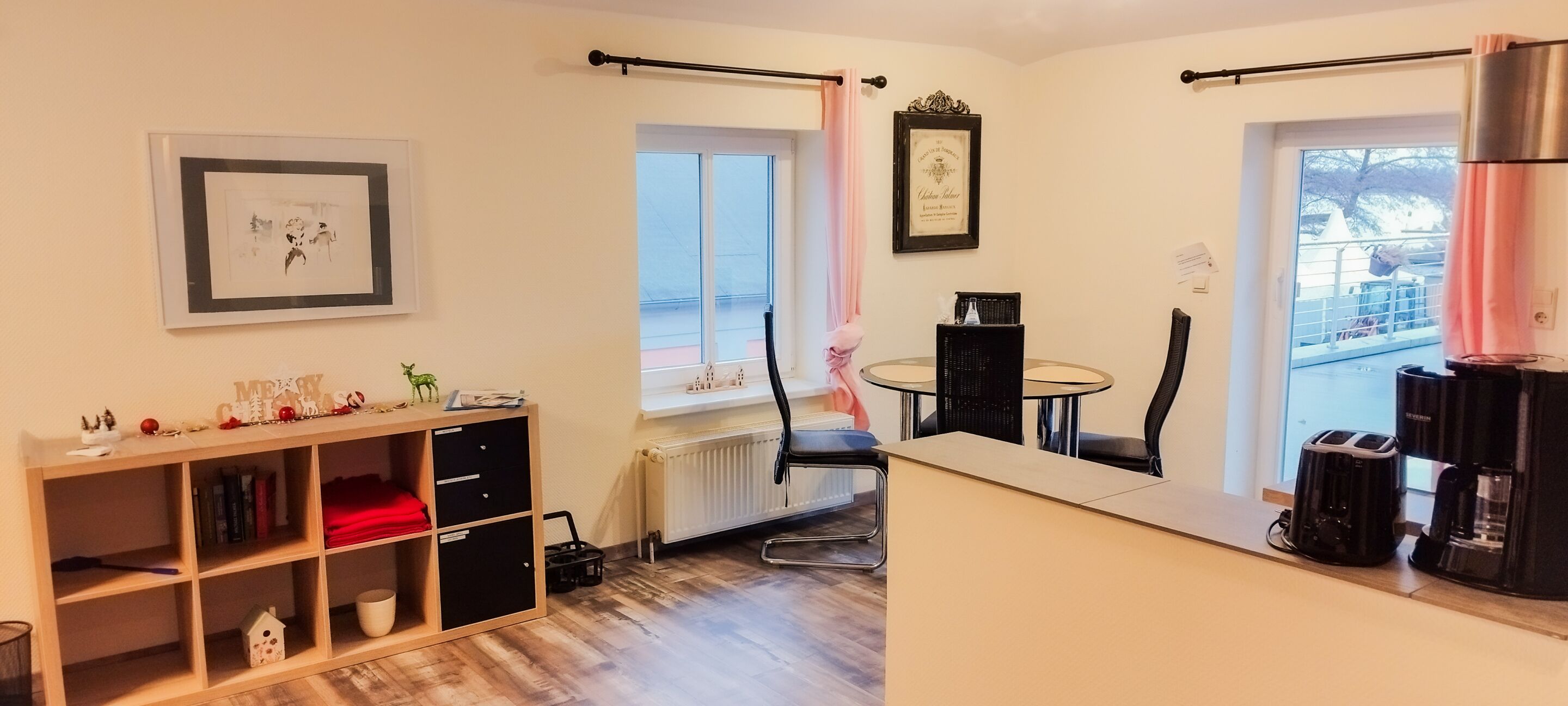 Una habitación con piso de madera, una estantería, una mesa de comedor con sillas, un radiador, una ventana con cortina y una cafetera.