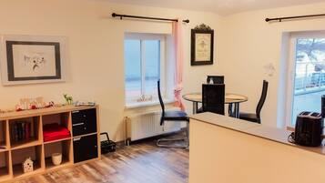 Una habitación con piso de madera, una estantería, una mesa de comedor con sillas, un radiador, una ventana con cortina y una cafetera.