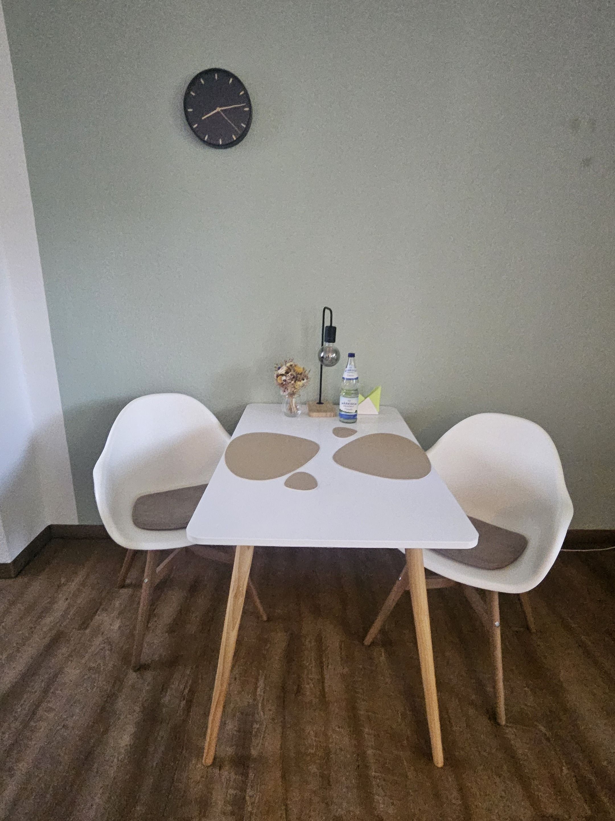 A small dining area with a white table, two white chairs, a clock on the wall, and a bottle of water on the table.