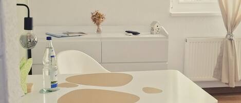 Un coin repas moderne avec une table blanche, une chaise blanche, une bouteille d’eau et une petite décoration représentant une maison, le tout posé sur la table.