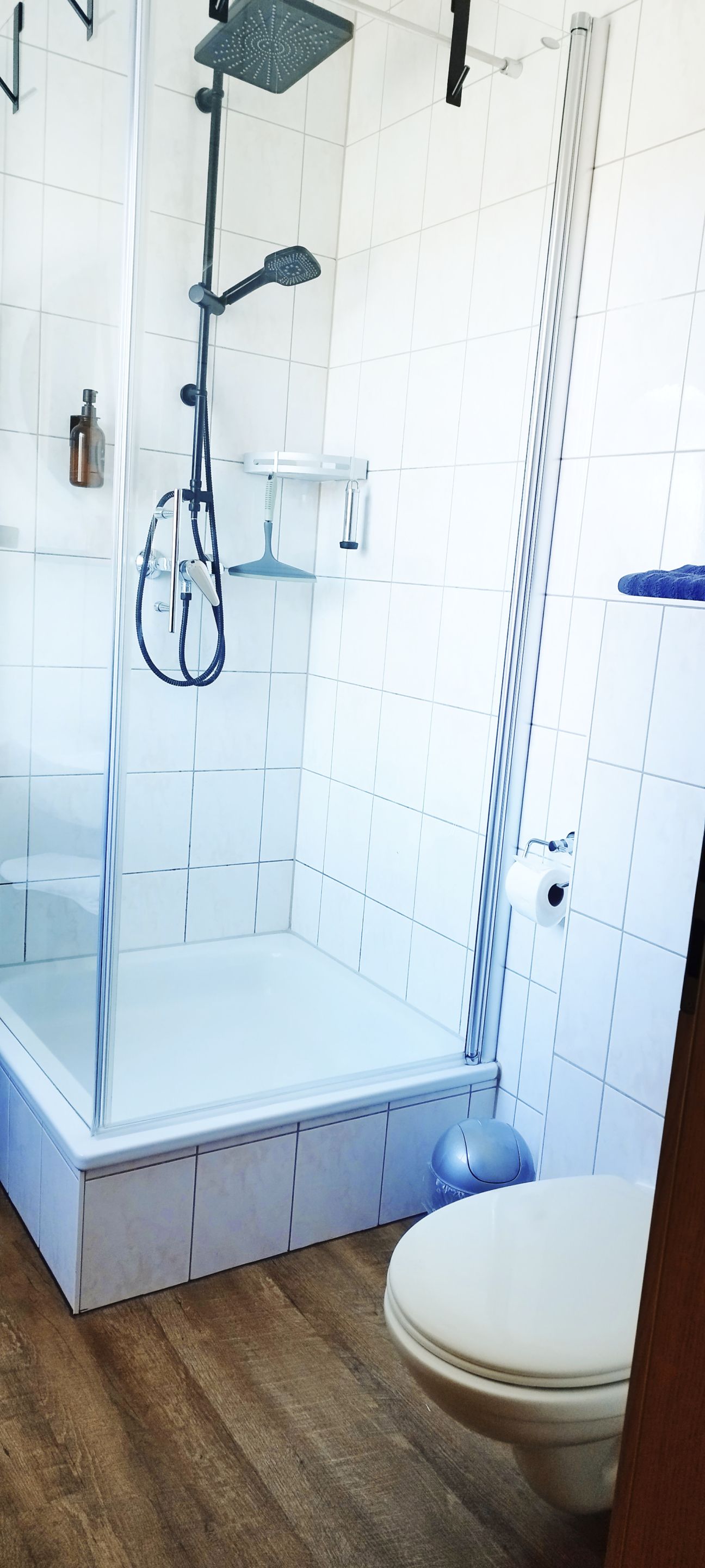 A modern bathroom with a glass-enclosed shower, white tiled walls, and a wooden floor.