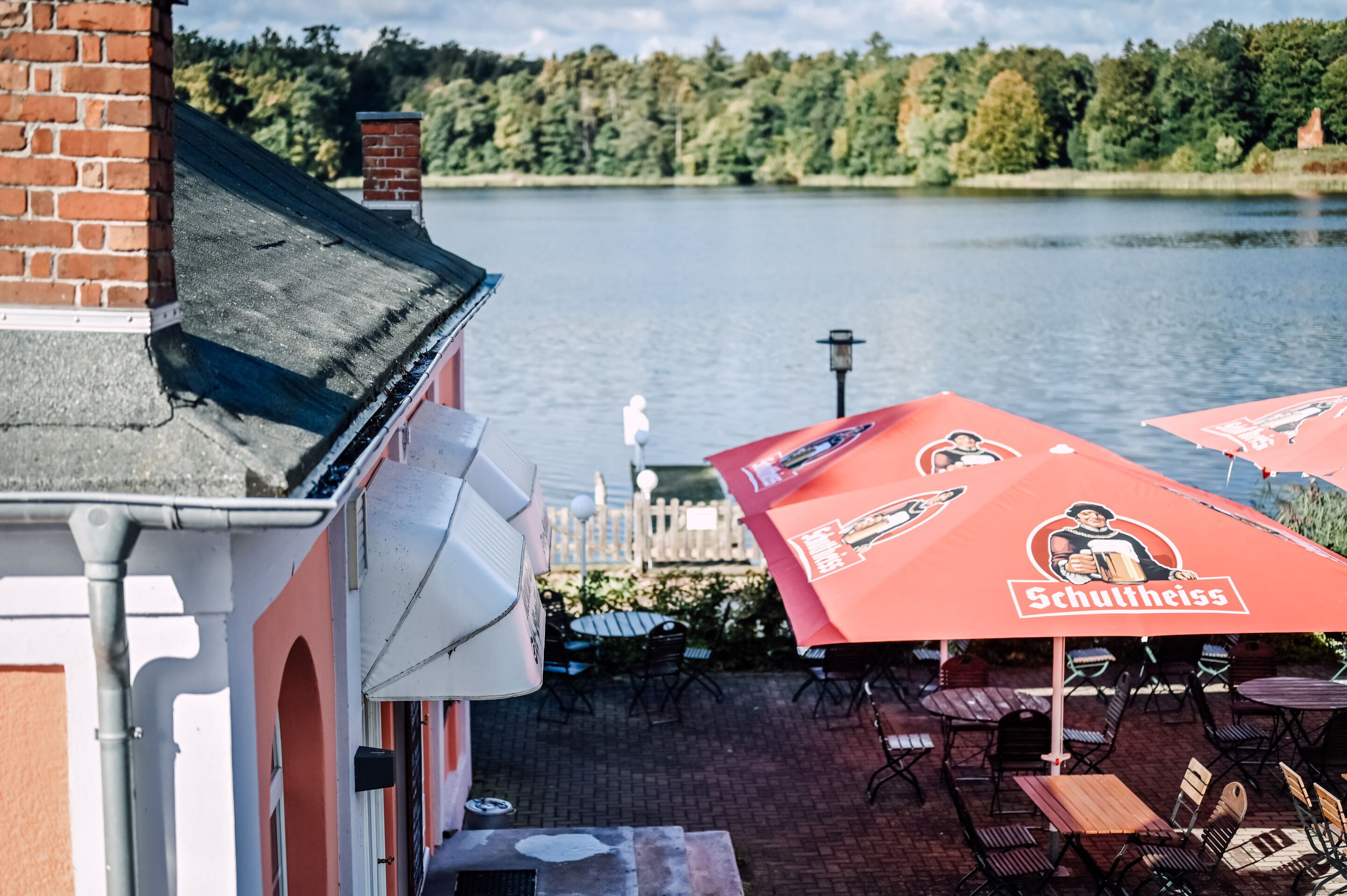 Un patio junto al lago con mobiliario exterior y una sombrilla roja con la marca 'Schultheiss'.
