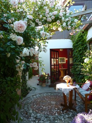 Un jardín con un porche de madera cubierto de rosales, un camino de ladrillo y una mesa con mantel blanco.