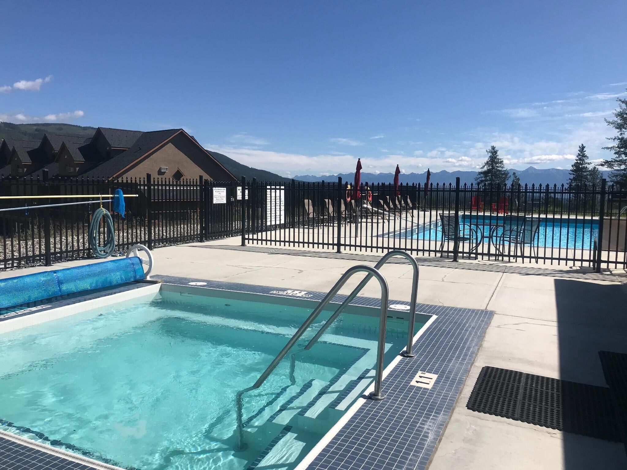 A swimming pool area with a fenced deck, a building in the background, and mountains in the distance.