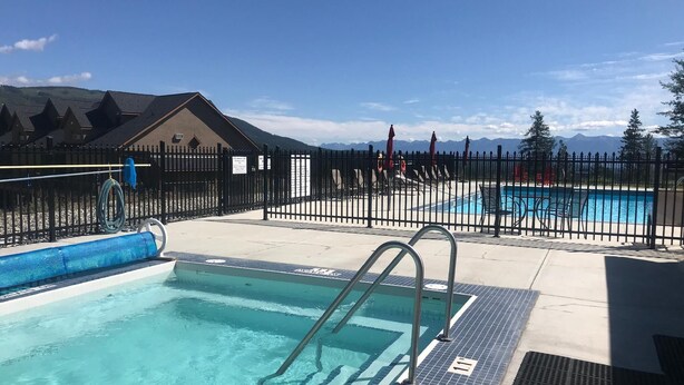 A swimming pool area with a fenced deck, a building in the background, and mountains in the distance. - Your clean, comfortable Rocky Mountain escape! True ski-in/ski-out 2BR condo right on the Main Run. Enjoy beautiful views, quality linens, a full kitchen, and year-round hot tub. The perfect, affordable base for family fun and relaxation. (Kimberley)