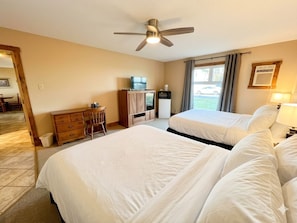 A hotel room with two beds, a desk with a chair, a ceiling fan, a window with curtains, and a view of the outside.