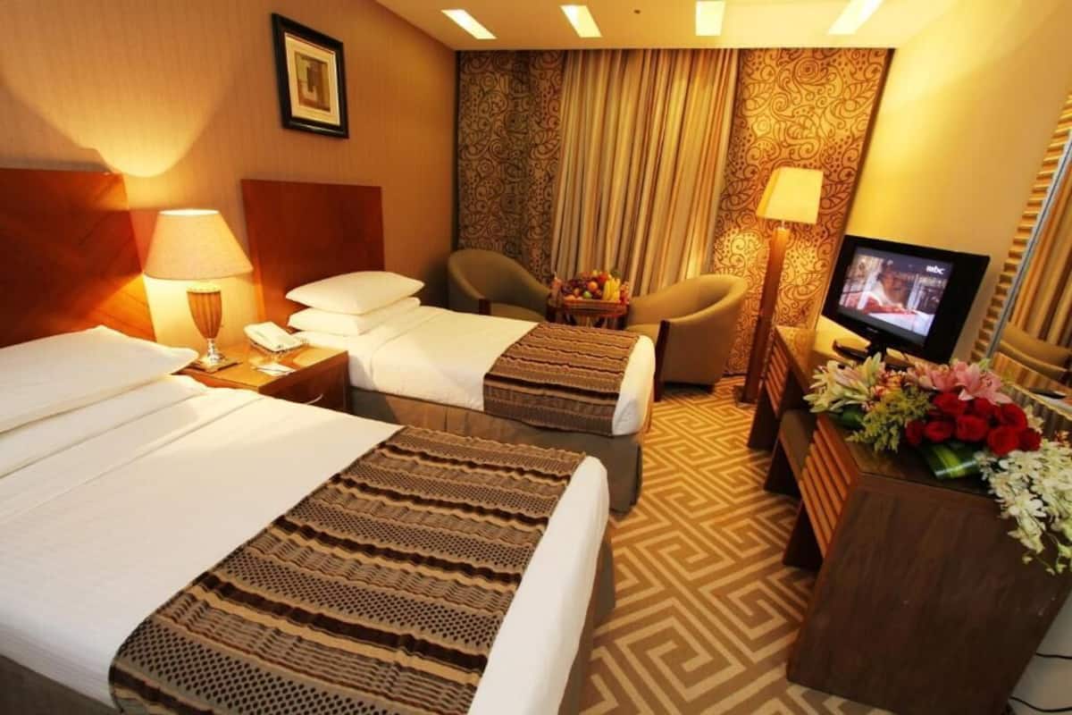 A hotel room with two beds, a television, a desk with a vase of flowers, and a patterned carpet.