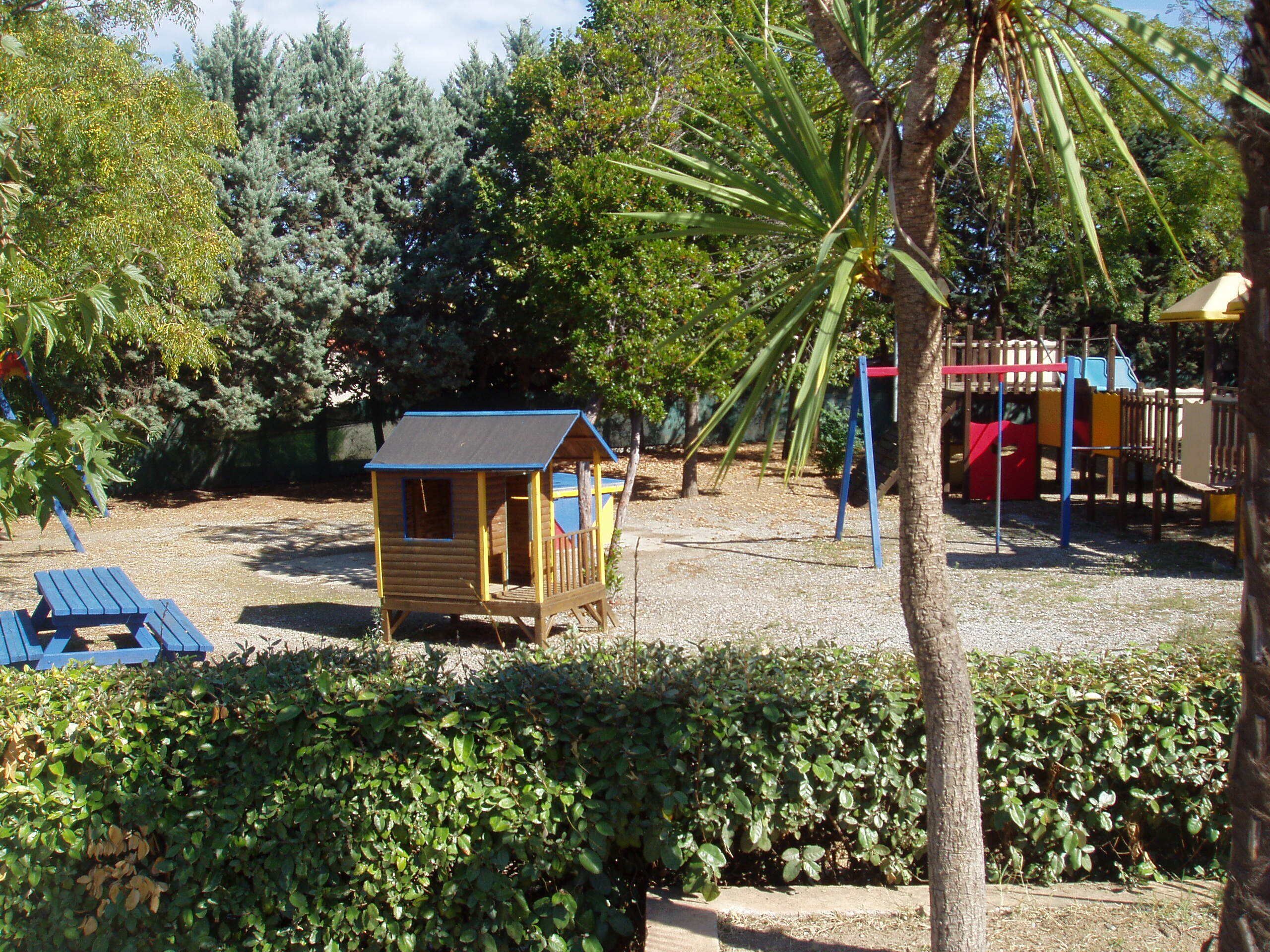 A wooden playhouse with a red roof, a small deck, and a blue bench in a park with trees and a playground in the background.