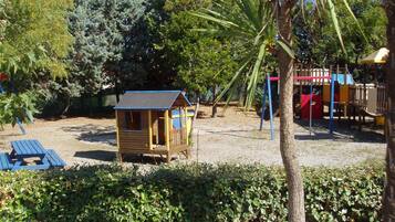 A wooden playhouse with a red roof, a small deck, and a blue bench in a park with trees and a playground in the background.