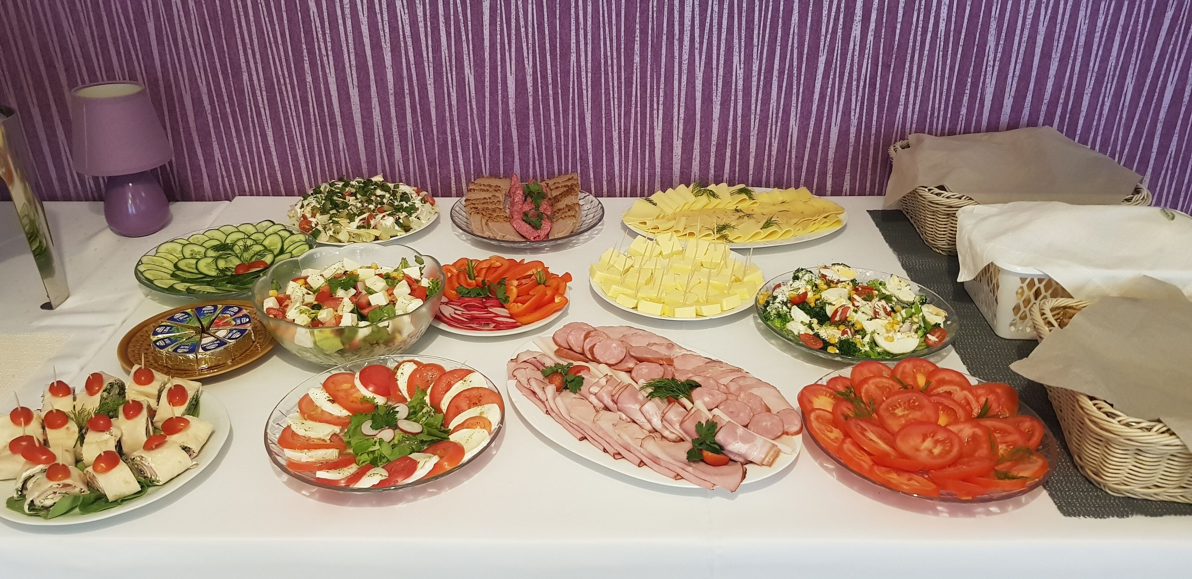 Una mesa con diversos alimentos, como ensaladas, sándwiches y fiambres, sobre un fondo a rayas morado.
