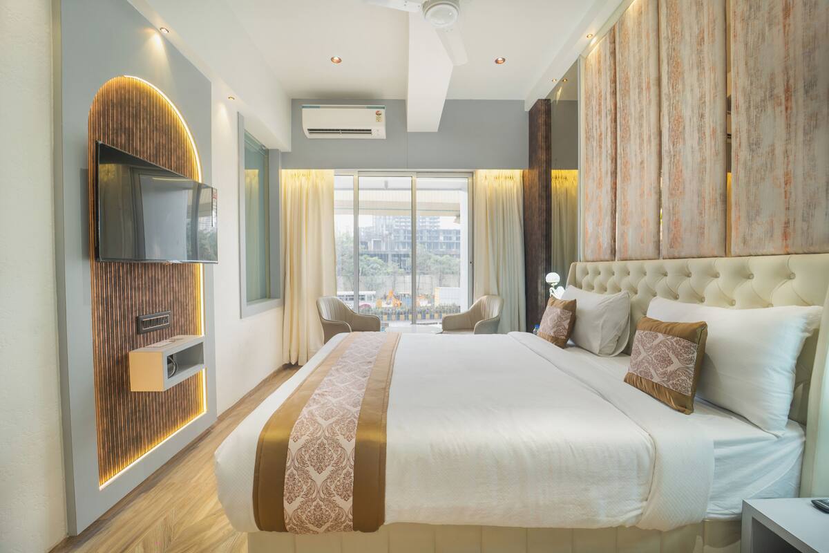 A modern hotel room with a large bed, a flat-screen TV mounted on the wall, a seating area with chairs, and a large window with curtains.
