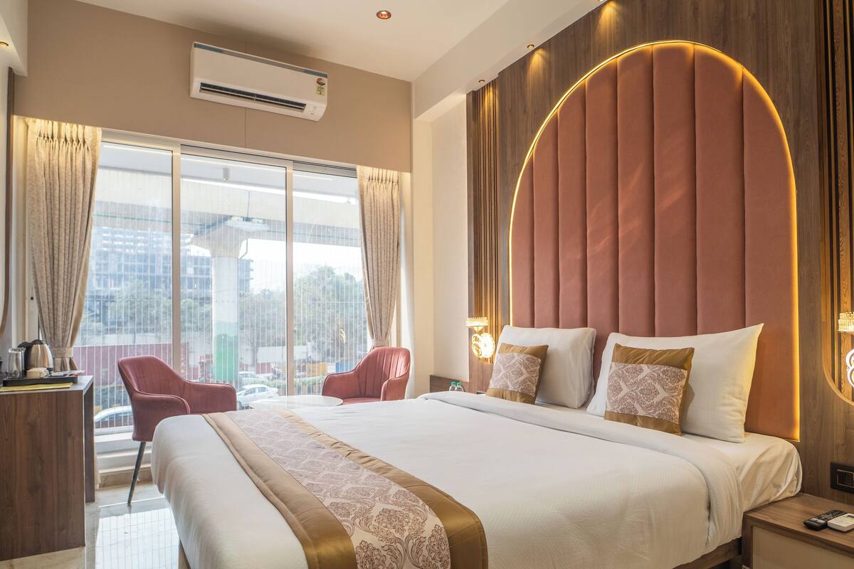 A modern hotel room with a large bed, a ceiling fan, an air conditioning unit, and a city view through large windows.