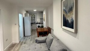 Egy modern nappali, szürke, moduláris kanapéval, étkezőasztallal, a háttérben pedig a konyha látható.