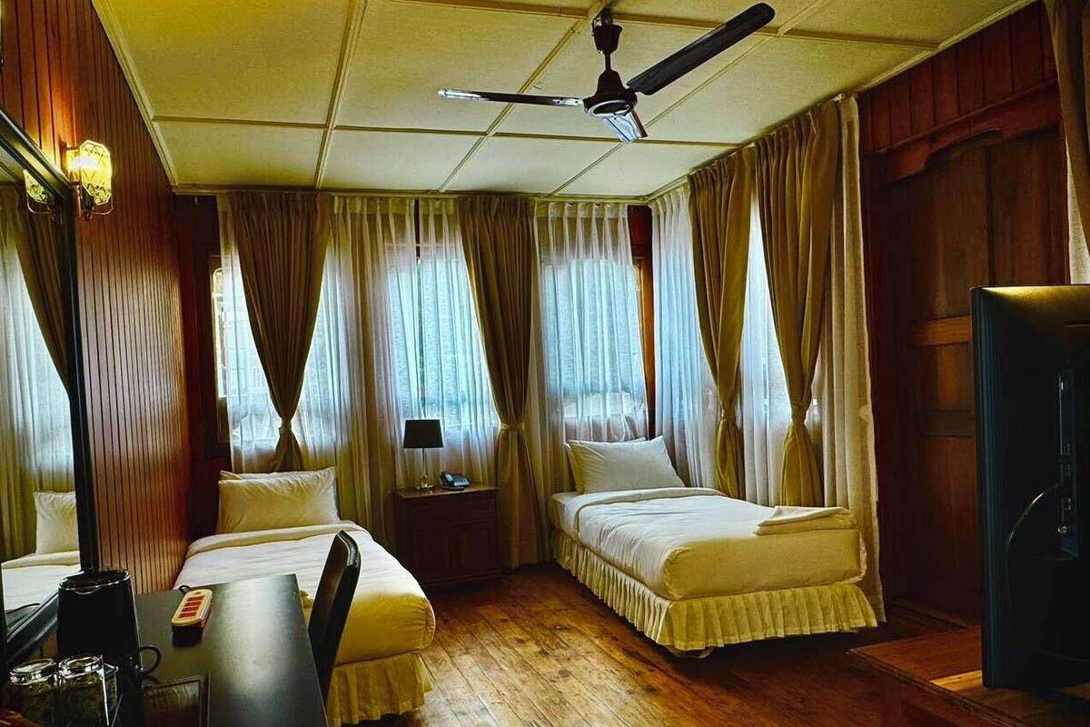 A hotel room with two beds, a desk with a lamp, a ceiling fan, and wooden flooring.