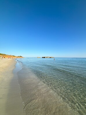 En strand med klart, grunt vatten, en sandstrand och avlägsna klippformationer.