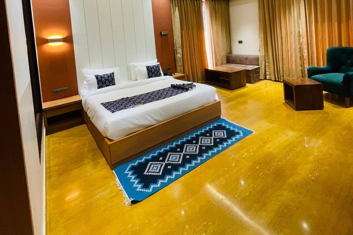 A modern hotel room with a bed, a ceiling fan, an air conditioning unit, a seating area with a sofa and armchairs, and a patterned rug.