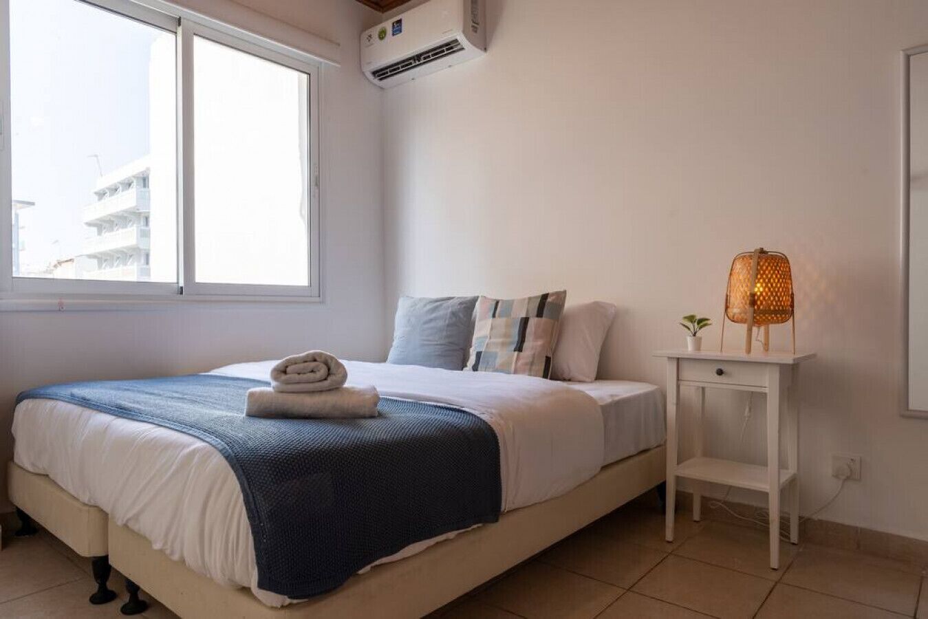 A neatly made bed with a blue blanket and a rolled towel, a bedside table with a lamp, and a window with an air conditioner.