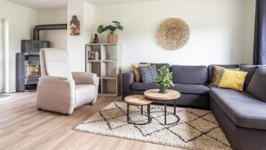 A modern living room with a beige armchair, a grey sofa, and a round coffee table. There is a bookshelf with books and a plant, and a decorative wall piece. - Hofvilla Winterswijk for 6 guests with 3 bedrooms and 2 bathrooms. Large garden, high comfort and pets welcome. (Kotten)