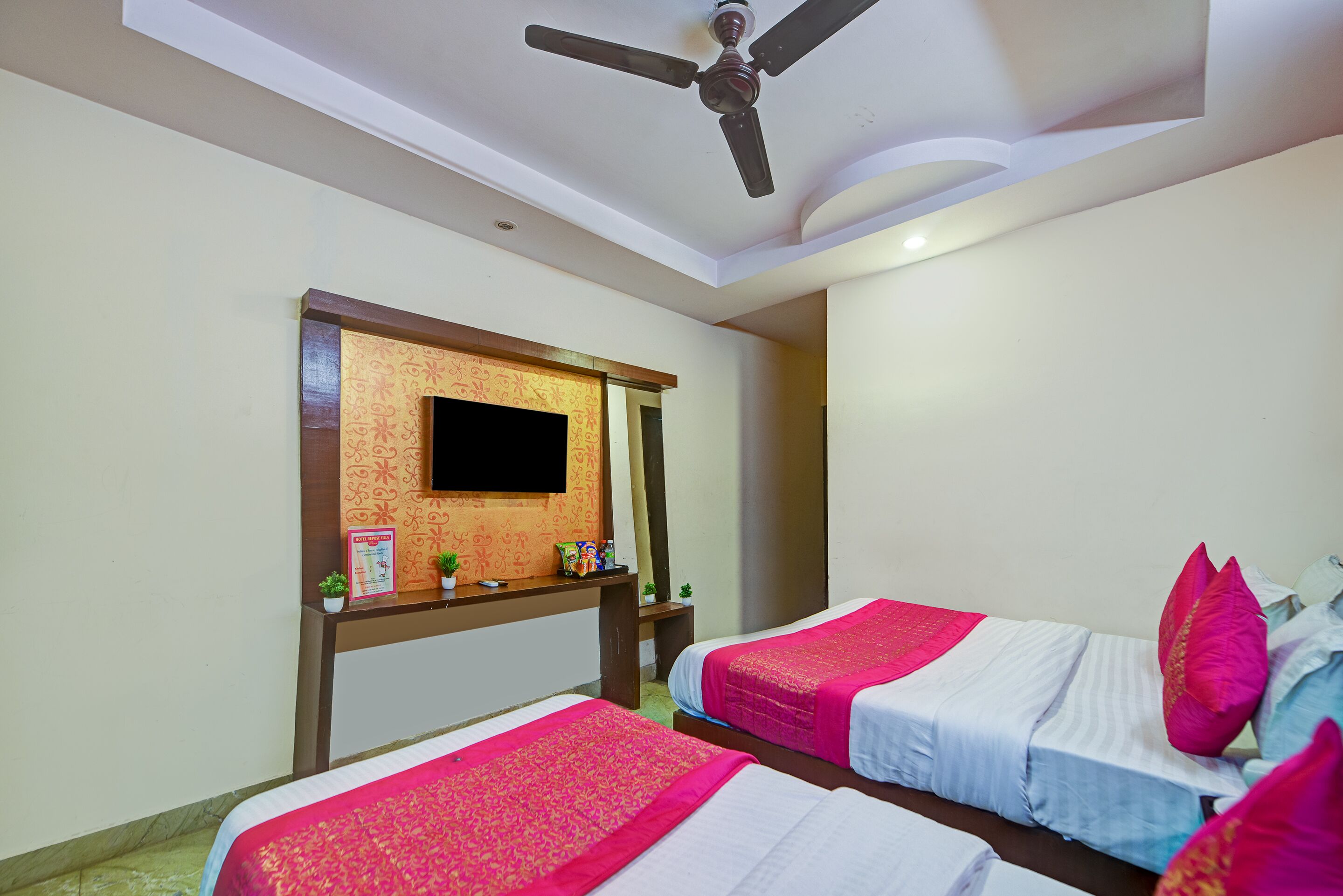 A hotel room with two beds, a TV mounted on a wooden panel, a ceiling fan, and a small table with decorative items.