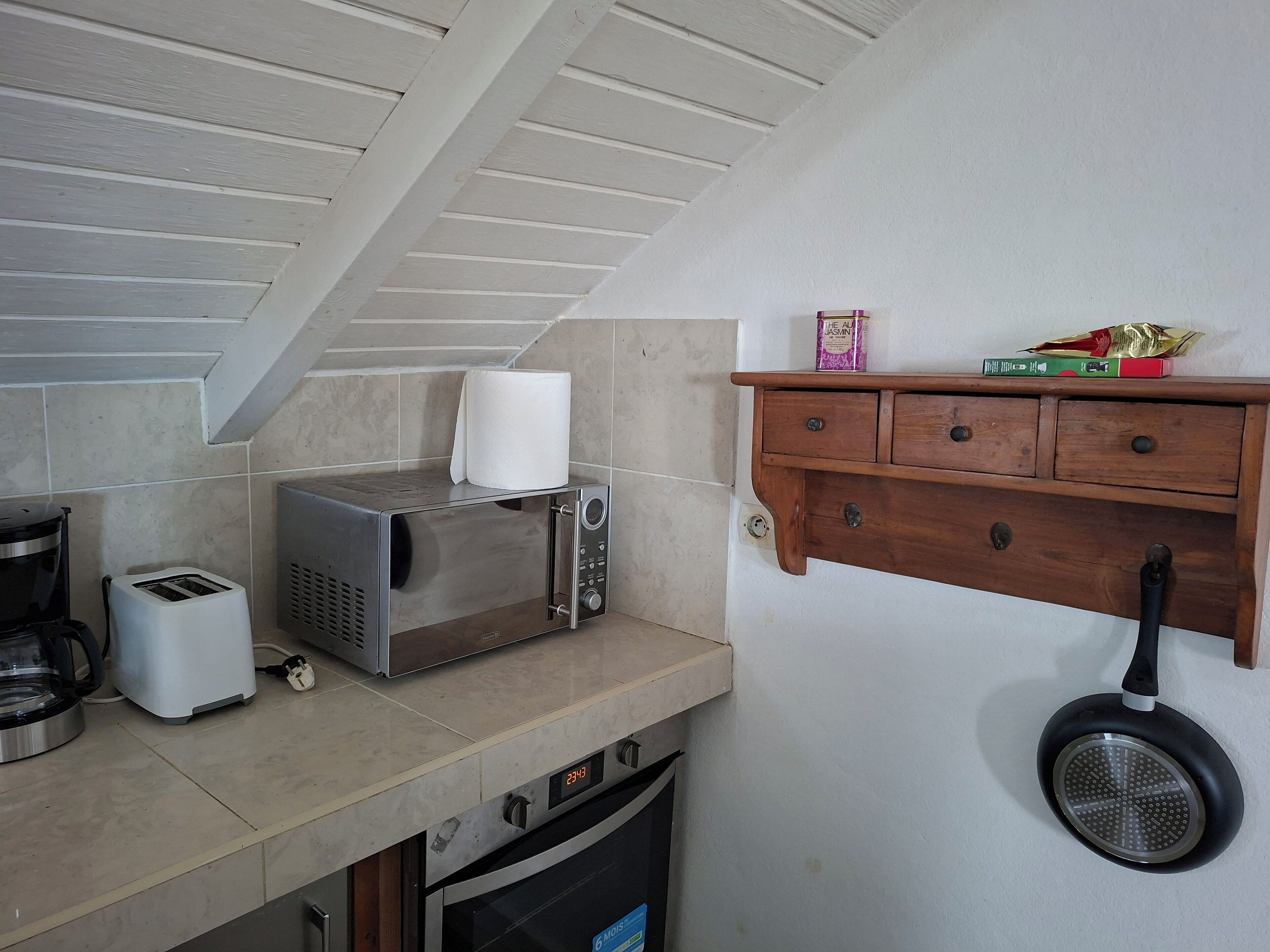 Une cuisine compacte équipée d’un grille-pain, d’un four à micro-ondes et d’un four traditionnel. Une étagère en bois contient quelques ustensiles et une poêle.