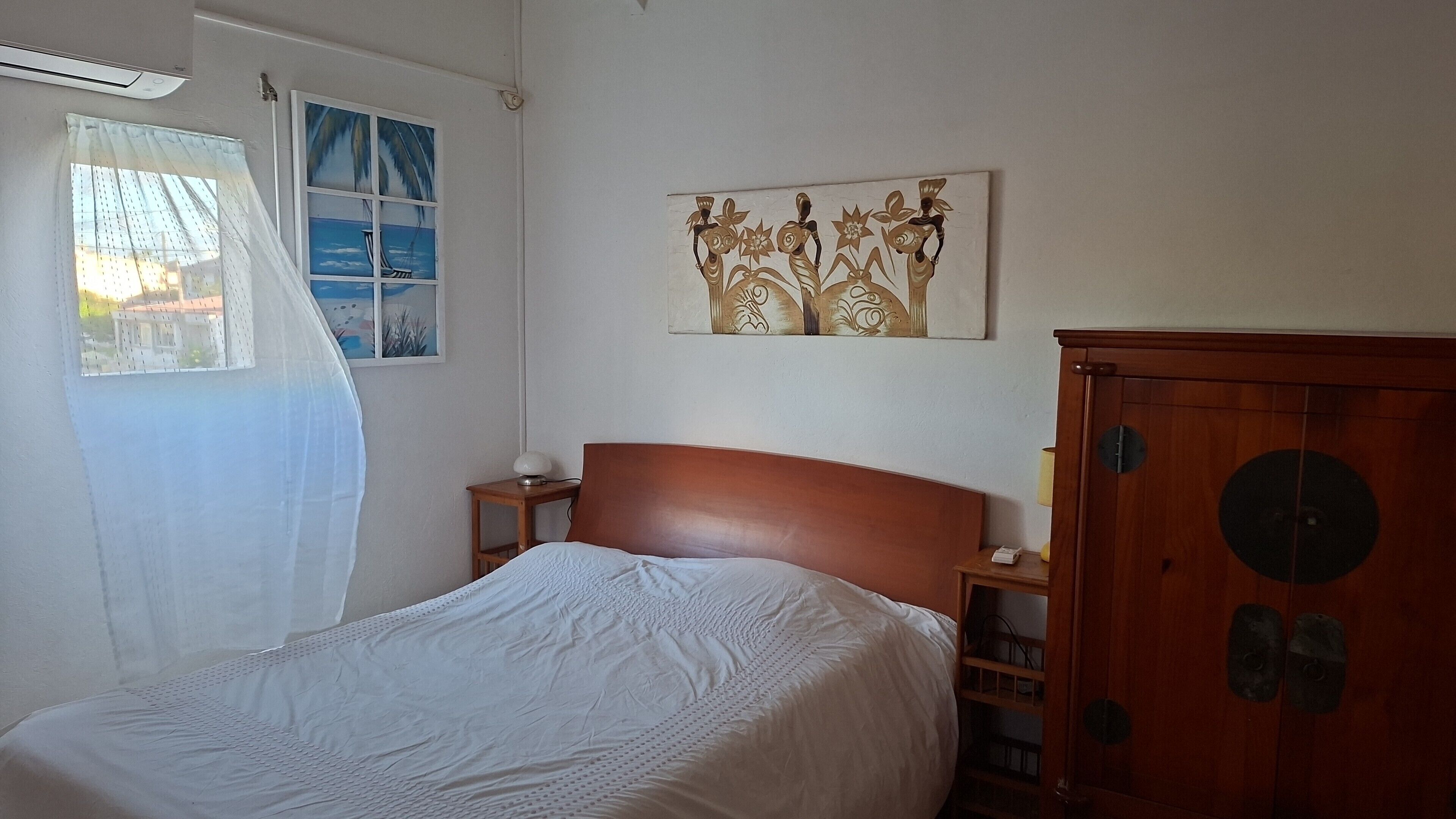 A bedroom with a bed, a wooden wardrobe, a nightstand, a wall-mounted painting, and a window with a view of a building.