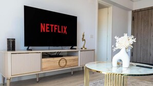 A modern living room with a large flat-screen TV displaying the Netflix logo, a wooden cabinet with drawers, a glass coffee table with gold legs, and a white vase with flowers. - Nest & Rest Brasov - Cozy Apartment (Brasov)