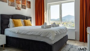 A bedroom with a large bed, a view of a cityscape through a window, and artwork on the wall. - Nest & Rest Brasov - Cozy Apartment (Brasov)