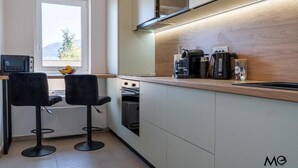 A modern kitchen with two bar stools, a microwave, a coffee maker, and a window with a view of greenery. - Nest & Rest Brasov - Cozy Apartment (Brasov)