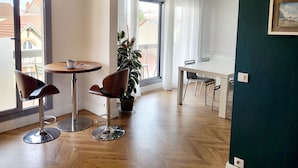 Una camera moderna con pavimento in legno, un tavolo rotondo con due sedie, una pianta in vaso e vista sugli edifici attraverso ampie finestre.