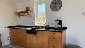 A modern kitchen with wooden cabinets, a black countertop, a coffee machine, and a clock on the wall. - Will's Ranch - Apartment (Den Helder)