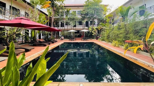 Une piscine entourée de verdure, agrémentée de chaises longues et de parasols, et bordée d’un bâtiment doté de balcons.