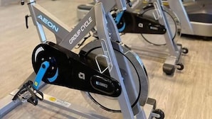 Un vélo d’appartement de la marque « AEON » et « GROUP CYCLE », placé dans une salle de sport dont le mur est orné d’un logo représentant un tigre.