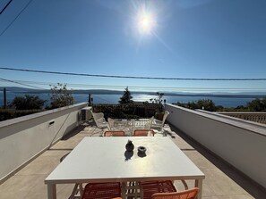 Eine Terrasse mit weißem Tisch und Stühlen, mit Blick auf ein Gewässer und ferne Berge.
