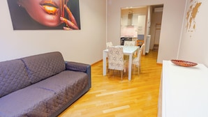 En stue med en grå sofa, et spisebord med mønstrede stole og et hvidt skab med en dekorativ skål.
